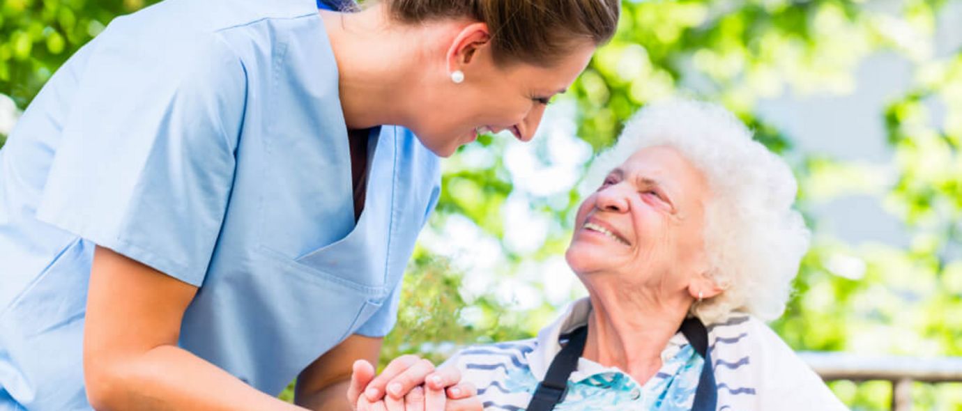 Altenpflegerin hält die Hand einer lächelnden Seniorin im Rollstuhl