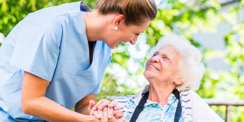 Altenpflegerin hält die Hand einer lächelnden Seniorin im Rollstuhl