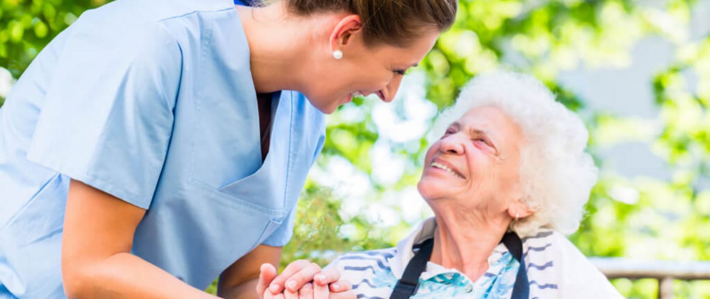 Altenpflegerin hält die Hand einer lächelnden Seniorin im Rollstuhl